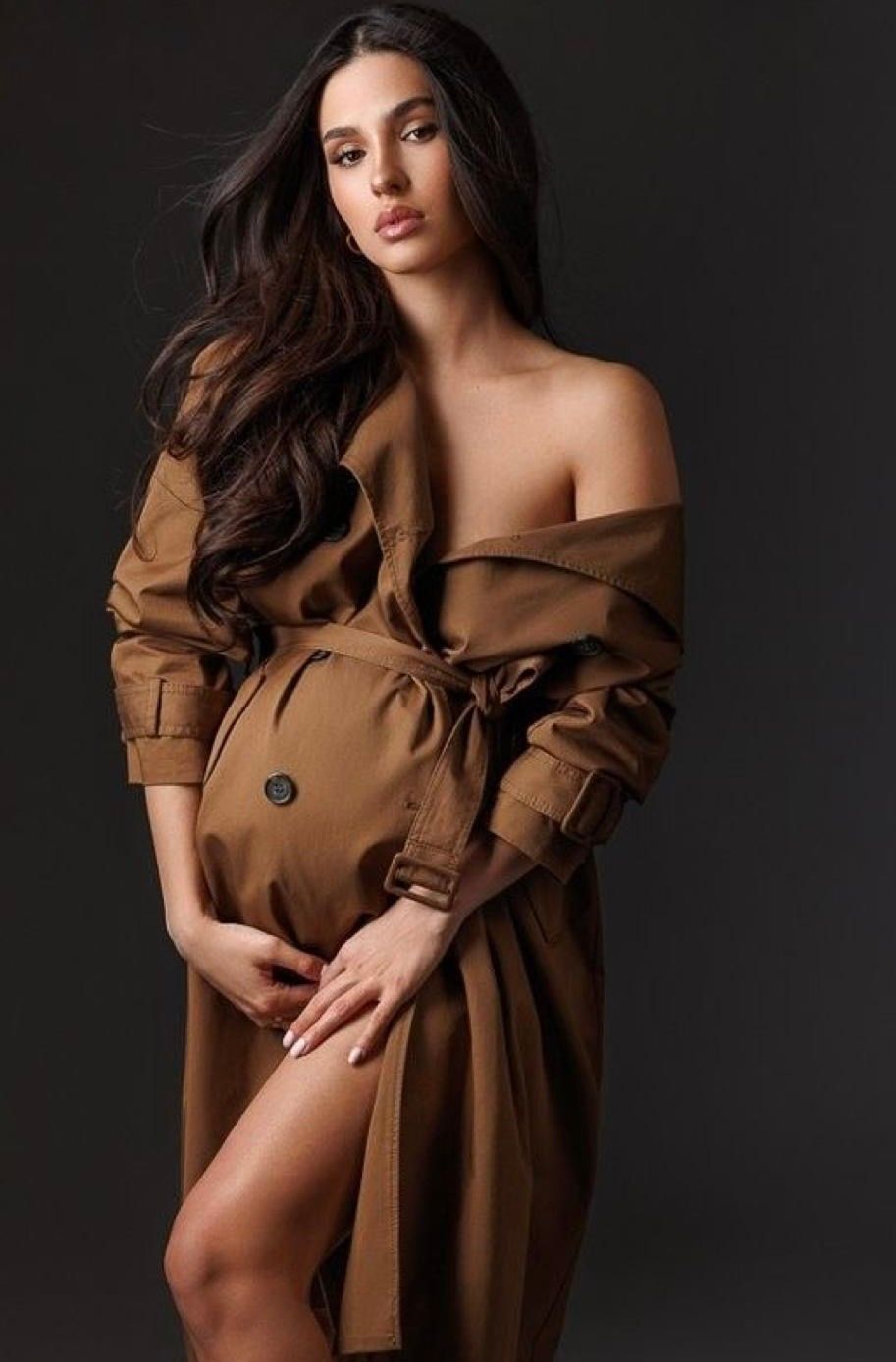 Pregnant woman in elegant off-shoulder camel trench coat posing gracefully, cradling baby bump in professional maternity photo.