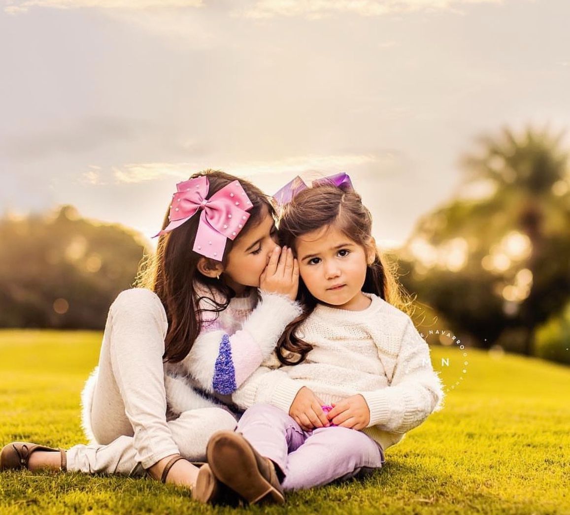 Two young girls with pink hair bows sit on green grass, one whispering in the other's ear at golden hour.