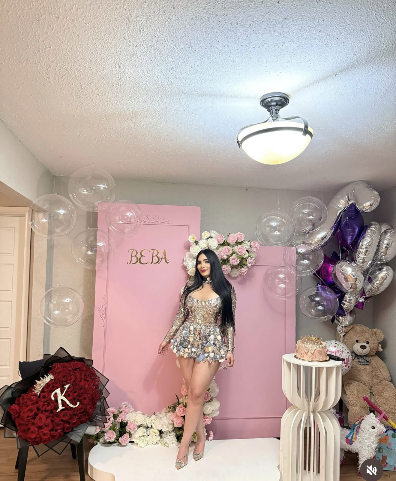 Woman in silver sequin mini dress poses at quinceañera celebration with pink backdrop and balloons.