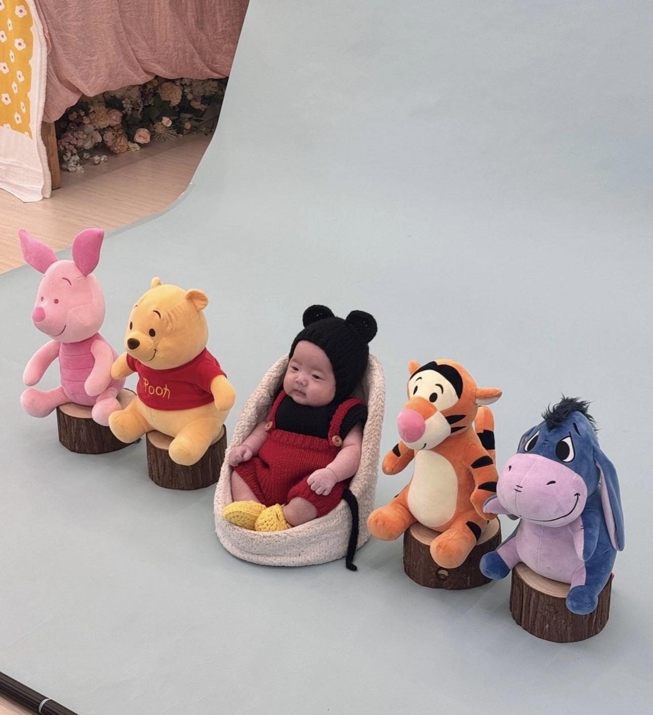 Baby dressed in bear costume sits in basket surrounded by Winnie the Pooh character plush toys on wooden stumps.