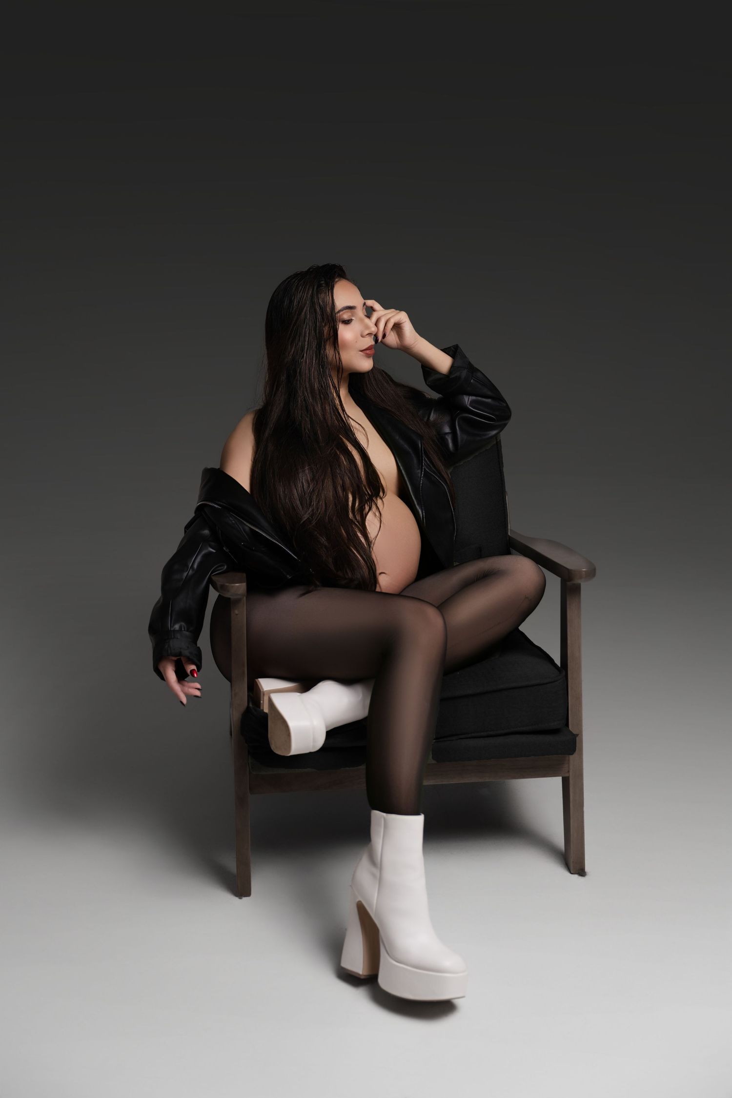Pregnant woman in black leather jacket, sheer tights, and white platform boots seated in a wooden chair on dark background.