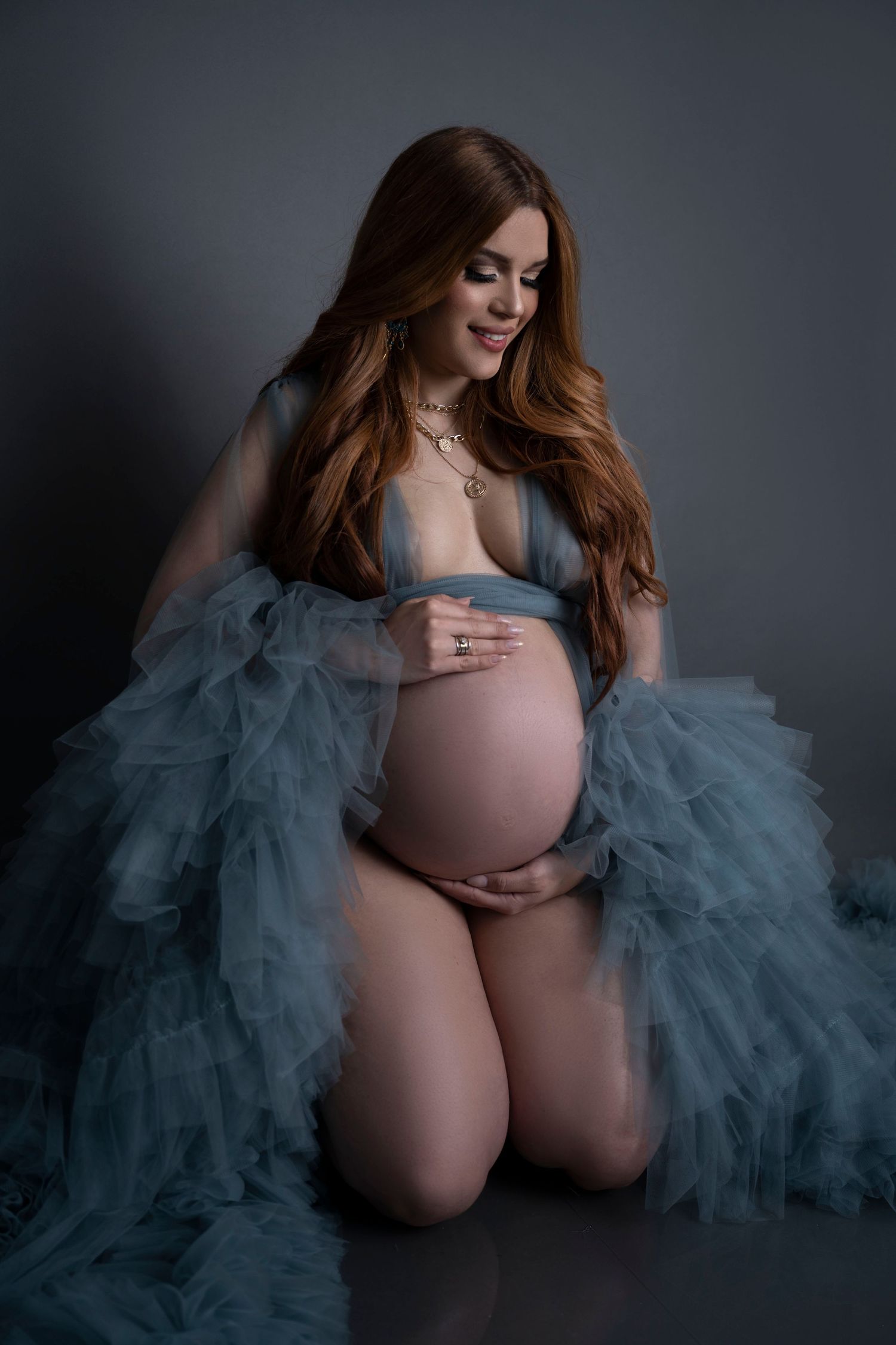 Pregnant woman kneeling in blue tulle robe, cradling baby bump, red hair, studio maternity portrait.