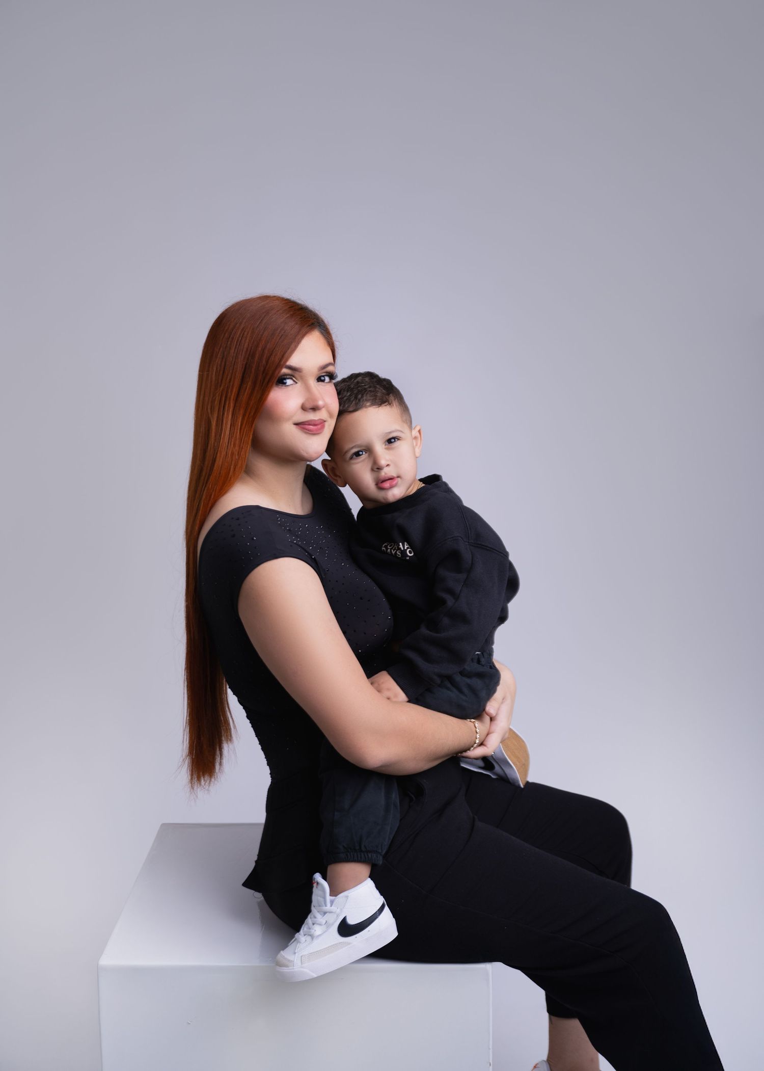 A young woman with red hair in black outfit sits on white cube holding a toddler boy in black Nike tracksuit.