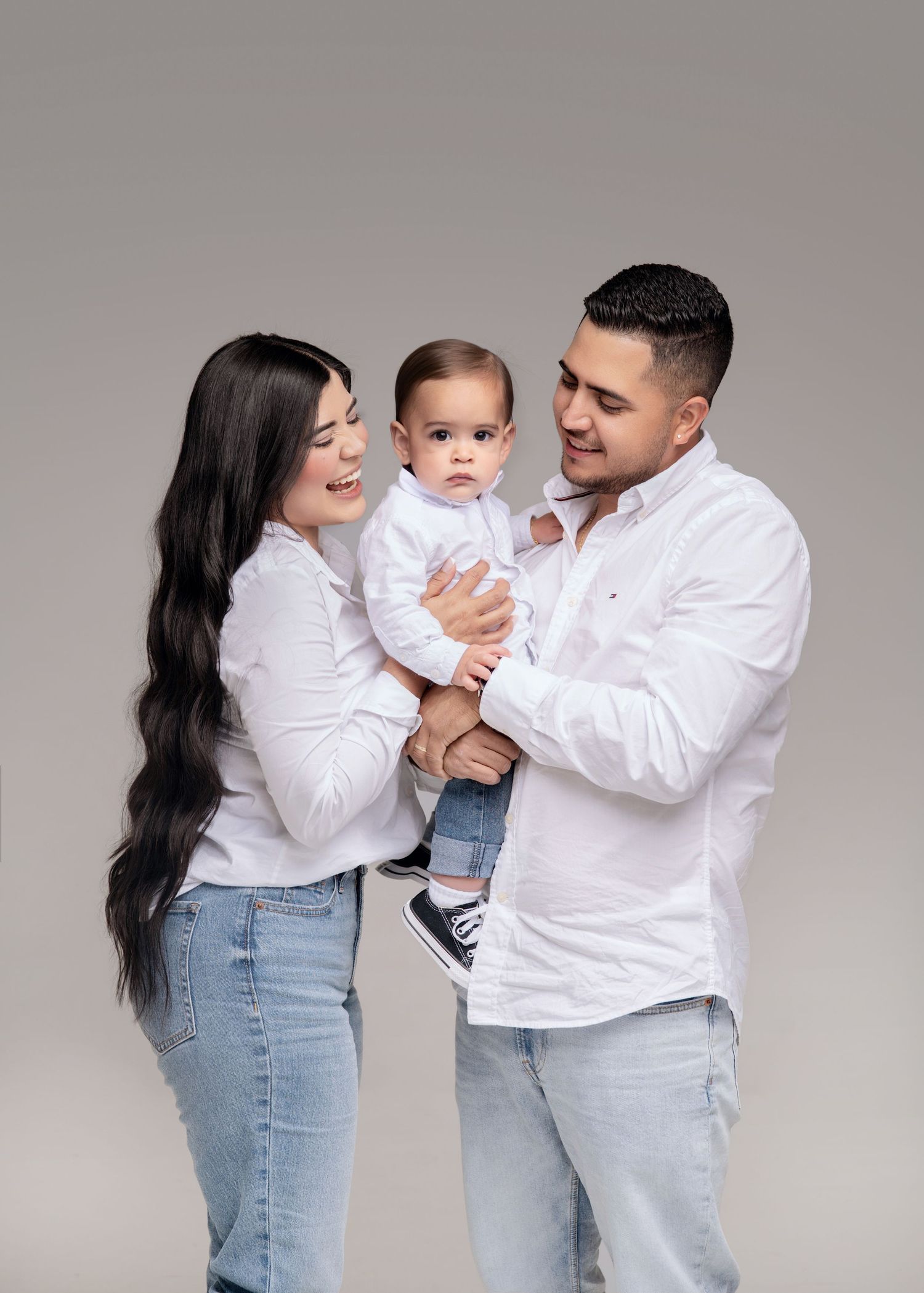 A young family in matching white shirts and jeans poses in a studio, parents smiling at their baby boy.