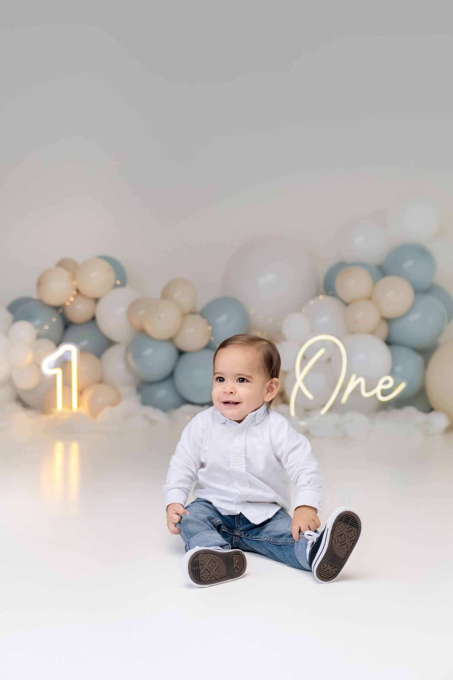 Smiling baby boy in white shirt and jeans sitting before balloon garland and neon one year birthday signs.