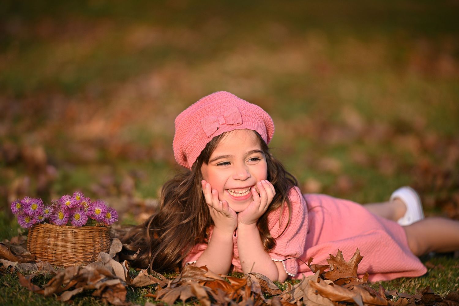 A person in pink attire lies on autumn leaves with a basket of flowers during a fall outdoor photography session.