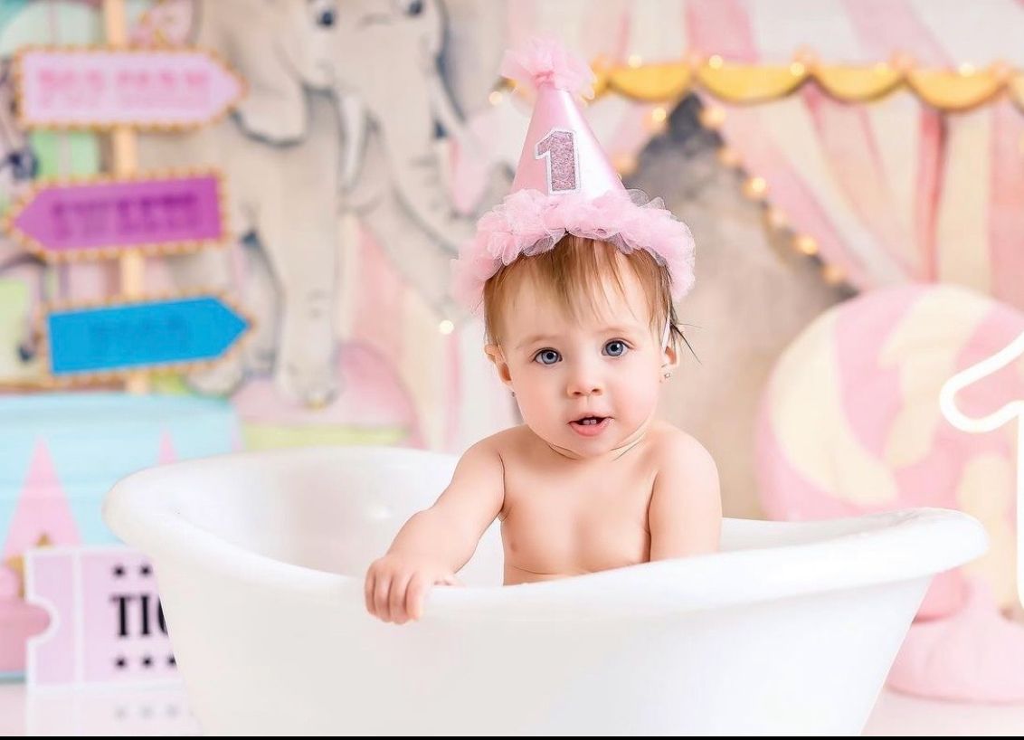 Baby wearing pink party hat sits in white bathtub with pink party decorations in background.