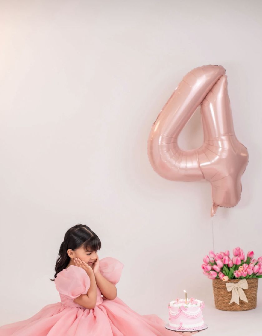 Pink birthday celebration setup with number 4 balloon, cake, flowers and tulle dress against white backdrop.