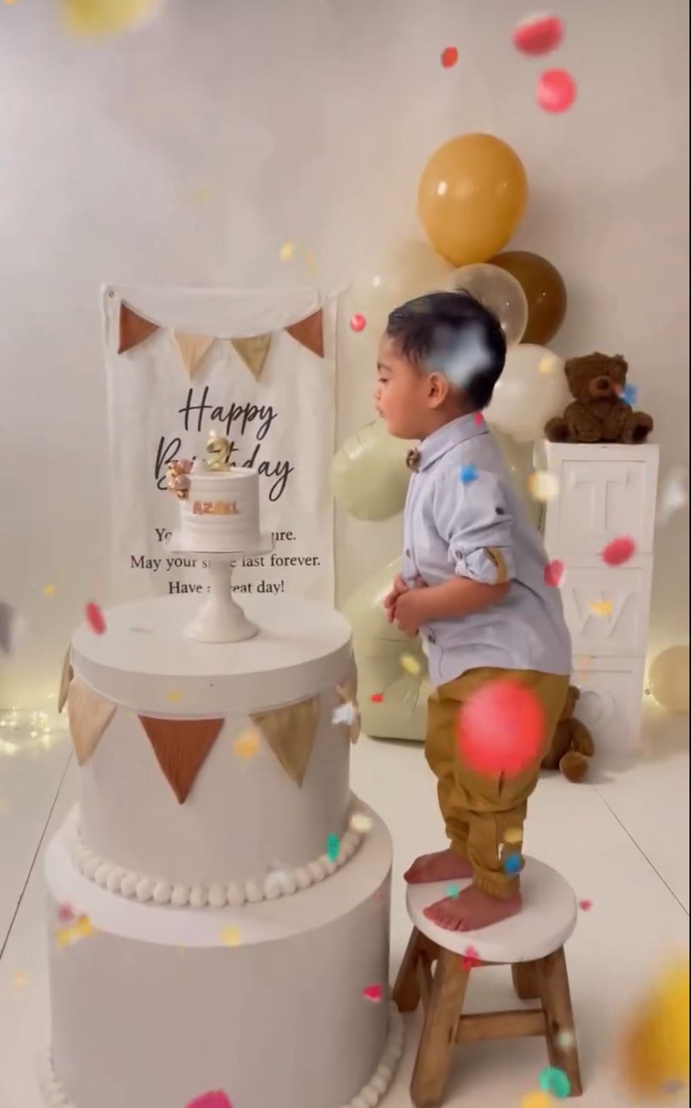 A minimalist two-tier white cake with bunting decorations and teddy bear displayed at a first birthday celebration.