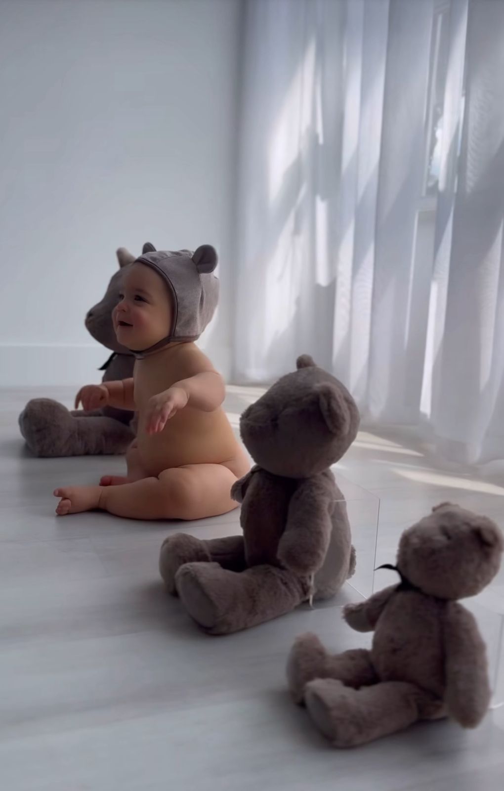 Baby and teddy bear playing together on white floor with natural light streaming through curtains.