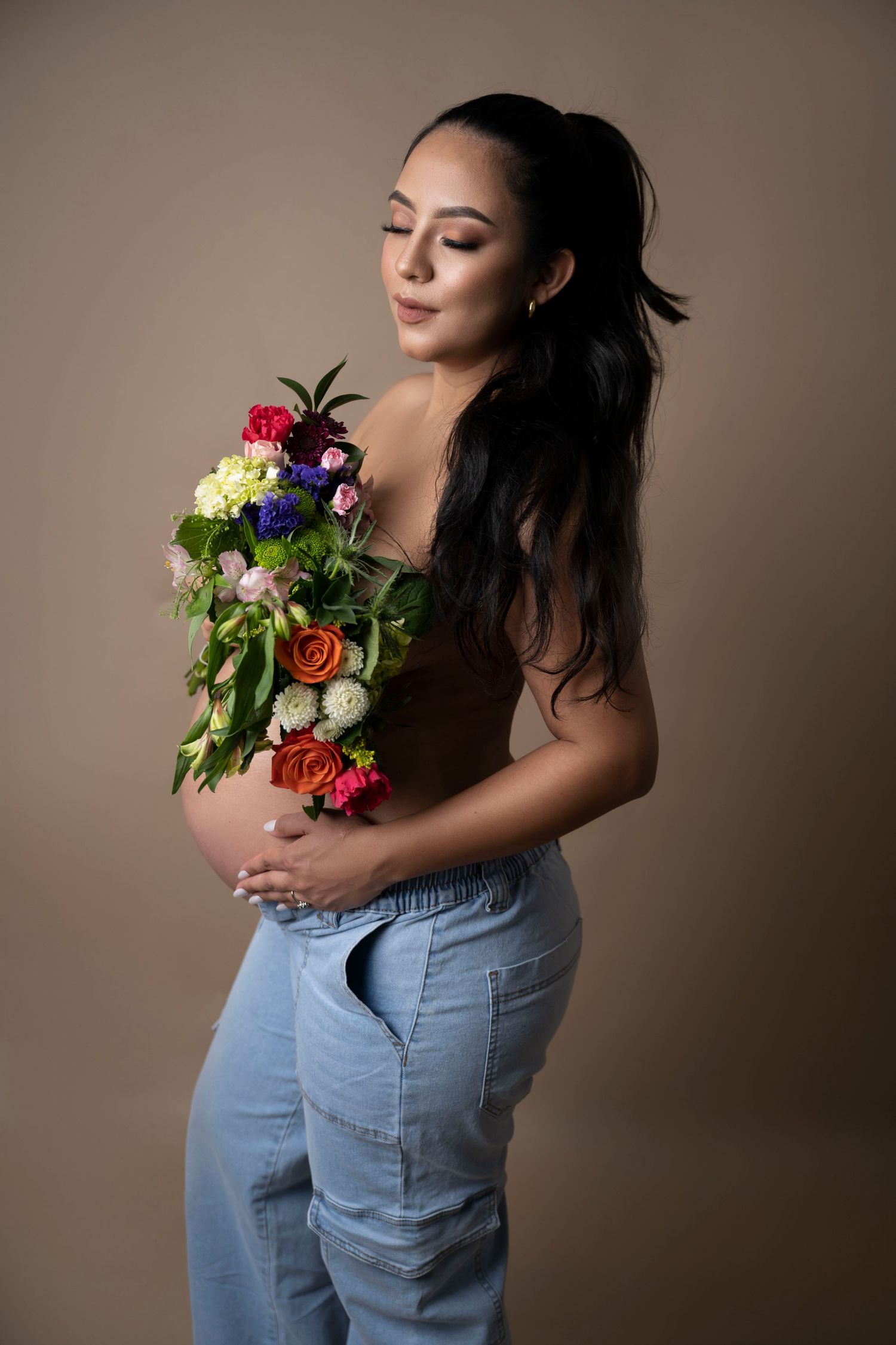 Expecting mother holding colorful bouquet wearing casual denim against neutral background.
