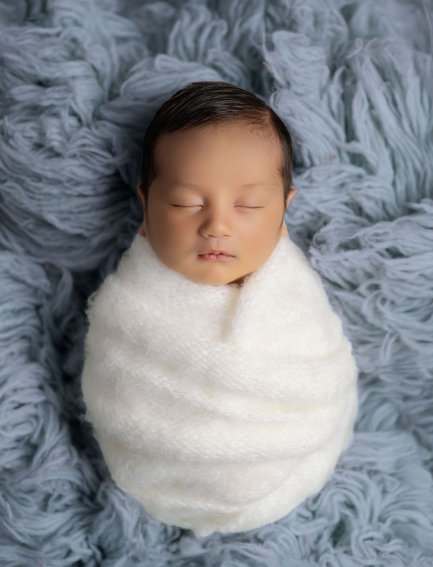 Sleeping newborn baby swaddled in white wrap on soft blue fuzzy blanket in professional studio photo.