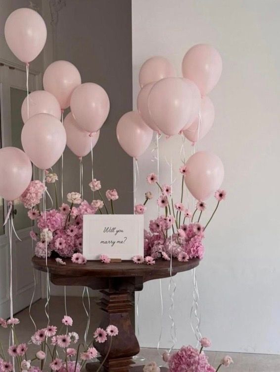 Romantic marriage proposal setup with pink balloons, flowers, and a will you marry me card on a wooden table.