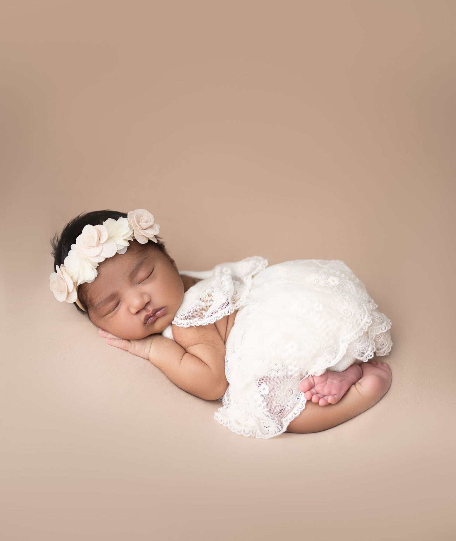 Sleeping newborn baby girl in white lace outfit and floral headband posed on beige backdrop.