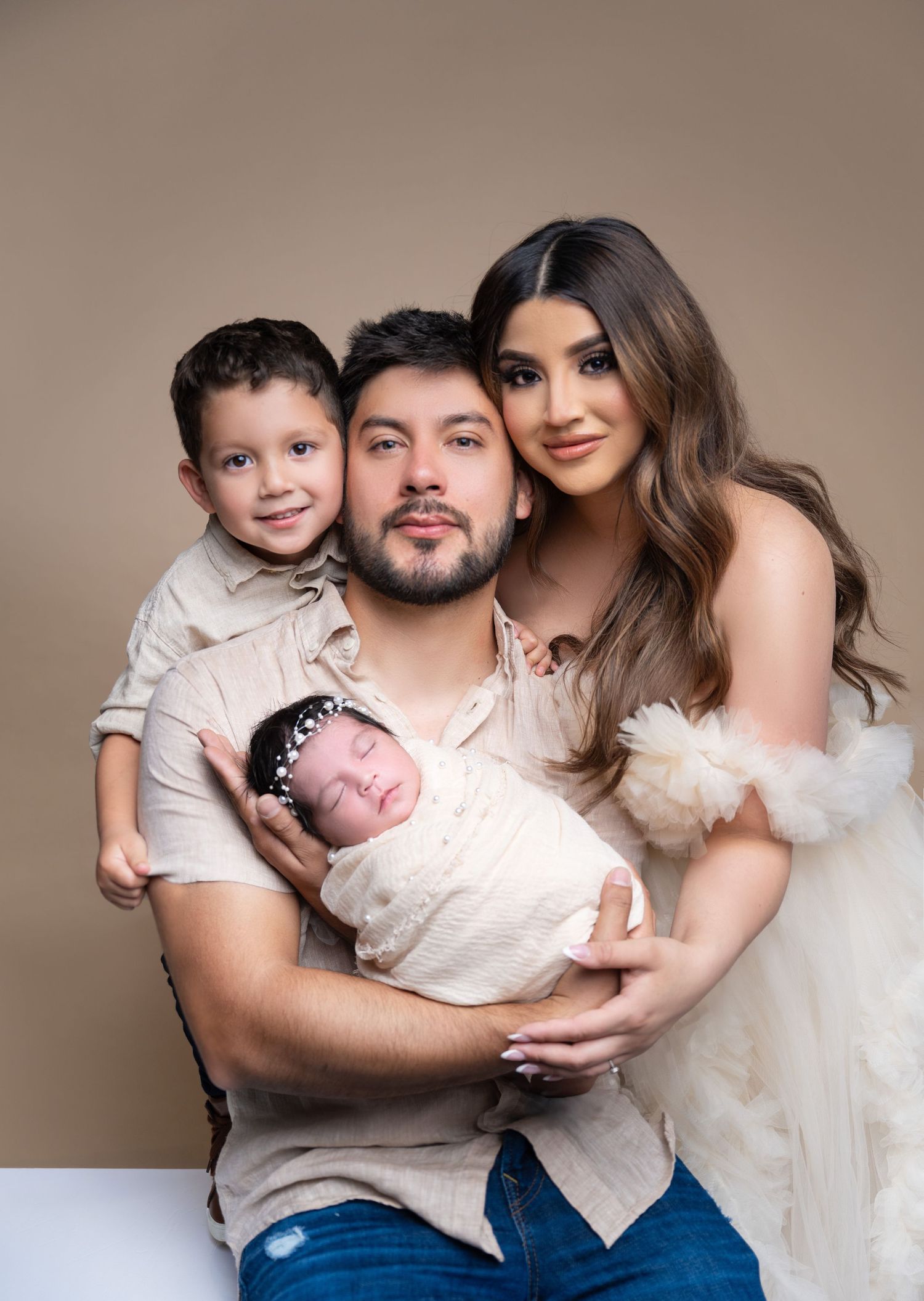 Family portrait with parents holding a toddler boy and swaddled newborn baby girl in a studio setting.