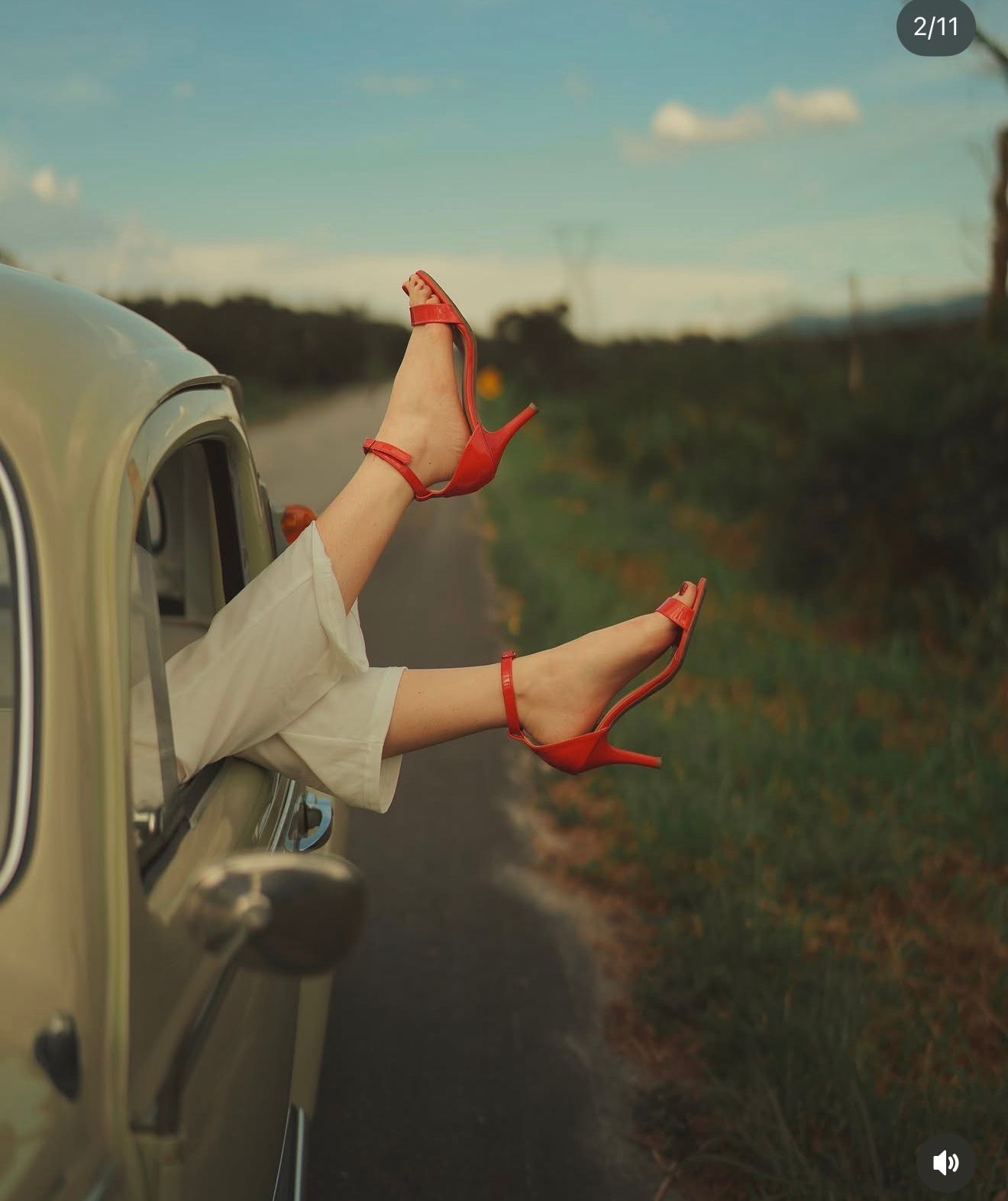 Woman's legs in red high heels dangling out of a vintage car window on a scenic country road.