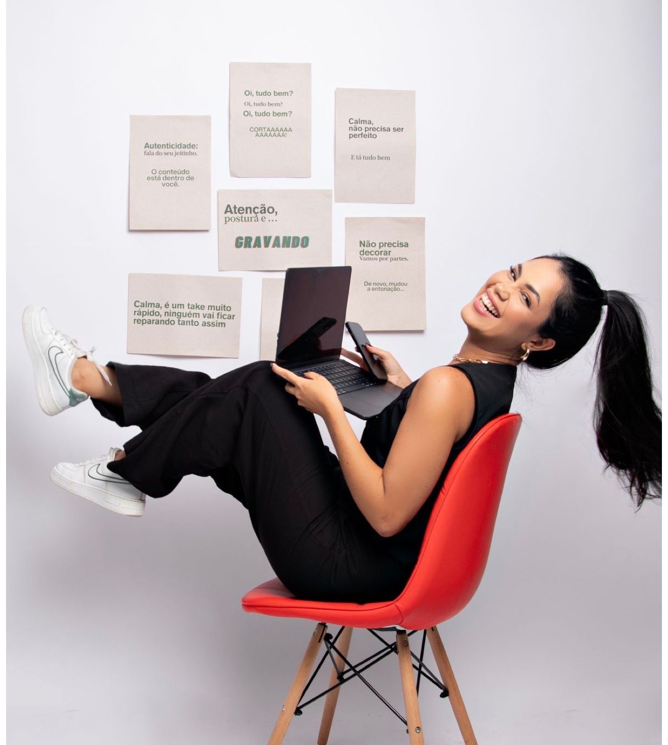 Young woman in black outfit sits on red chair holding laptop, smiling, with motivational Portuguese notes on wall.