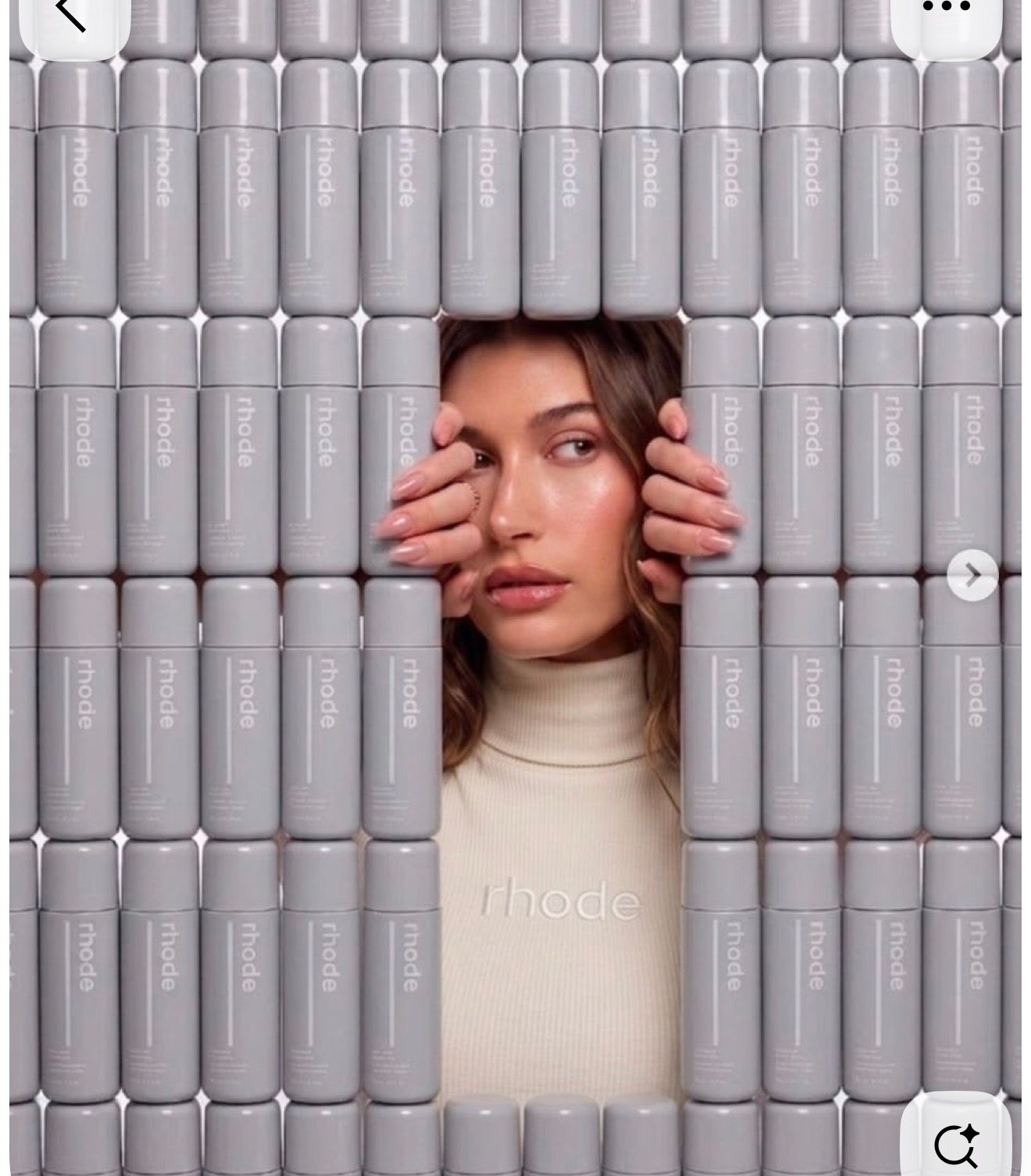 Woman peering through a wall of Rhode skincare lip treatment tubes arranged in rows, wearing a Rhode branded sweater.