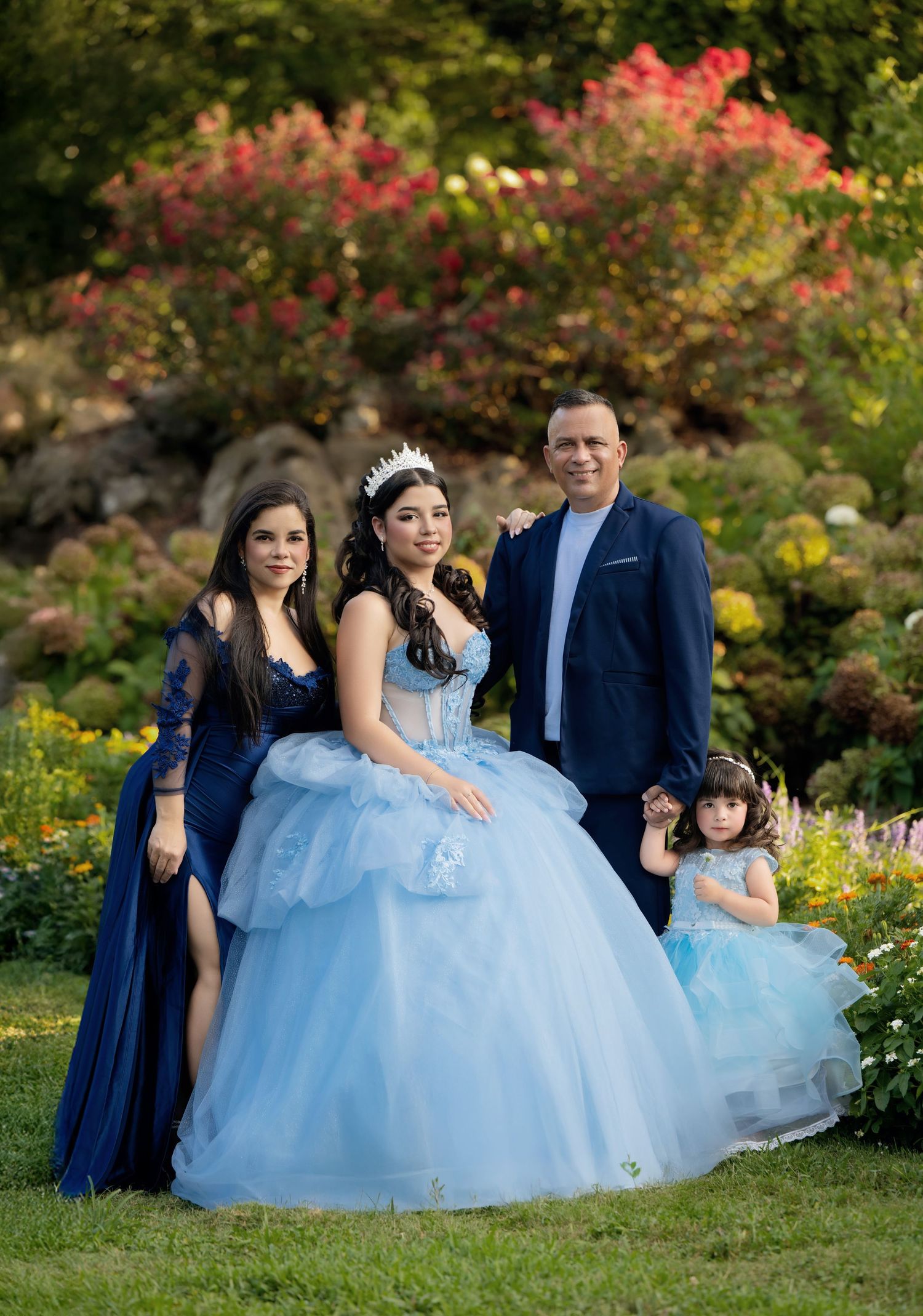 Family posing in garden for quinceañera celebration, girl in blue ball gown with tiara, surrounded by colorful flowers.