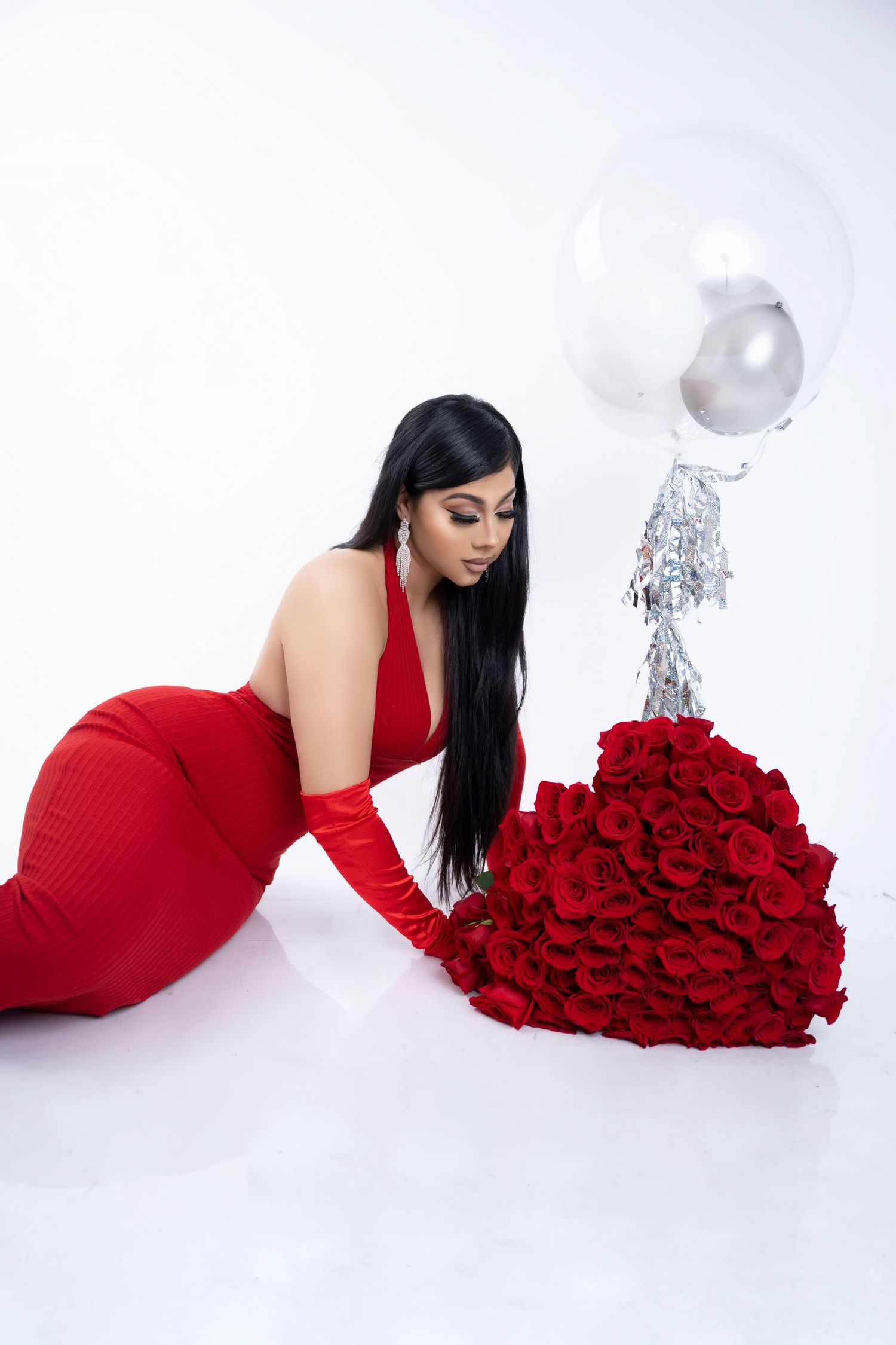 Woman in a red dress and gloves kneels beside a large bouquet of red roses with a silver balloon against a white studio backdrop.