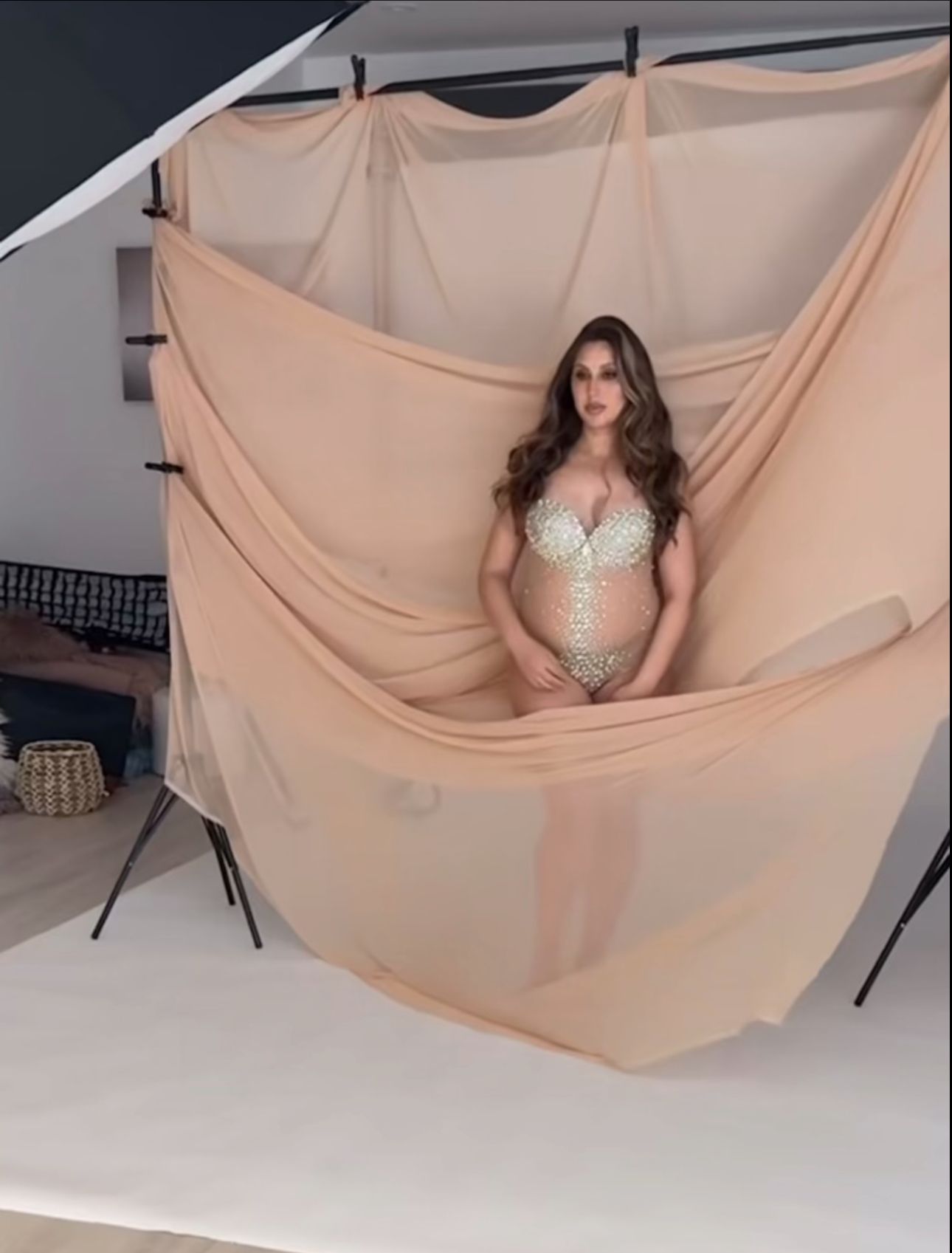 Woman in embellished bodysuit poses in front of peach draped fabric backdrop in a professional photo studio setting.