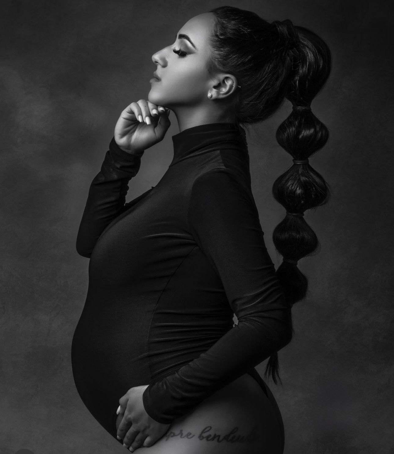 Black and white maternity photo of a pregnant woman in a turtleneck bodysuit with a bubble ponytail, posing elegantly.