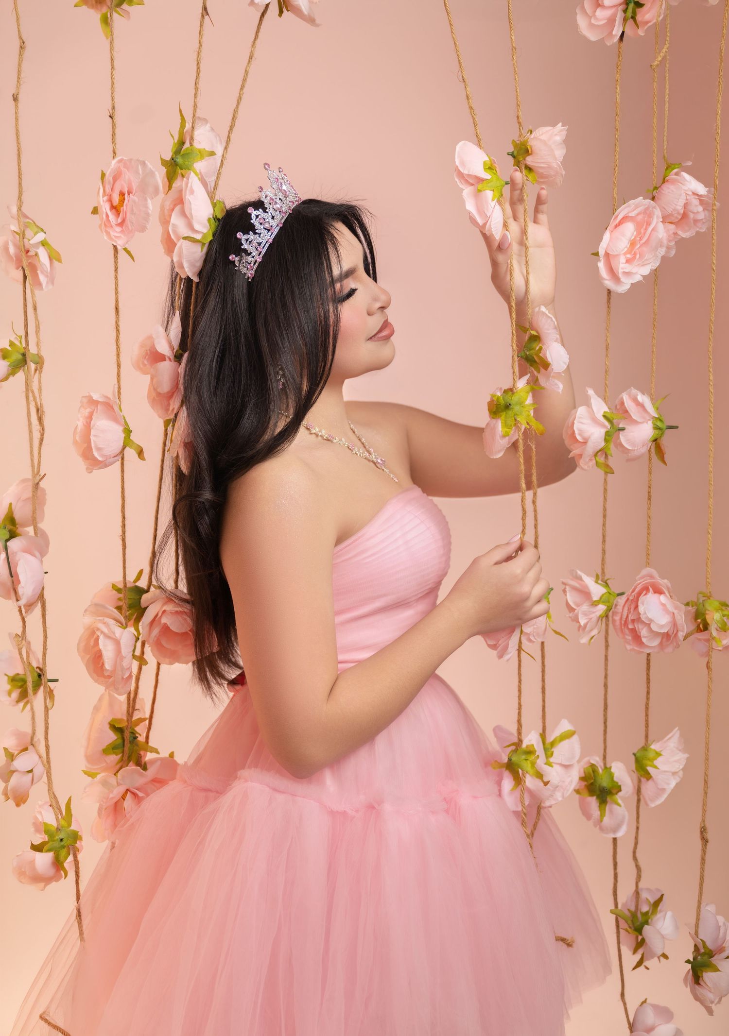 Young woman in pink tulle gown and tiara poses gracefully among hanging pink roses on a soft blush background.