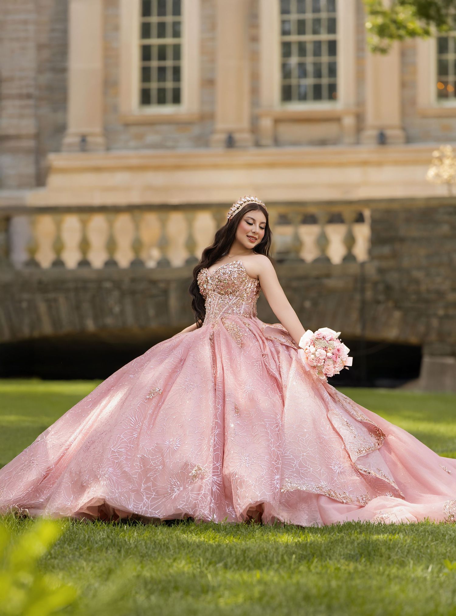 Young woman in pink ball gown with gold embroidery holds bouquet at elegant quinceañera photoshoot outdoors.