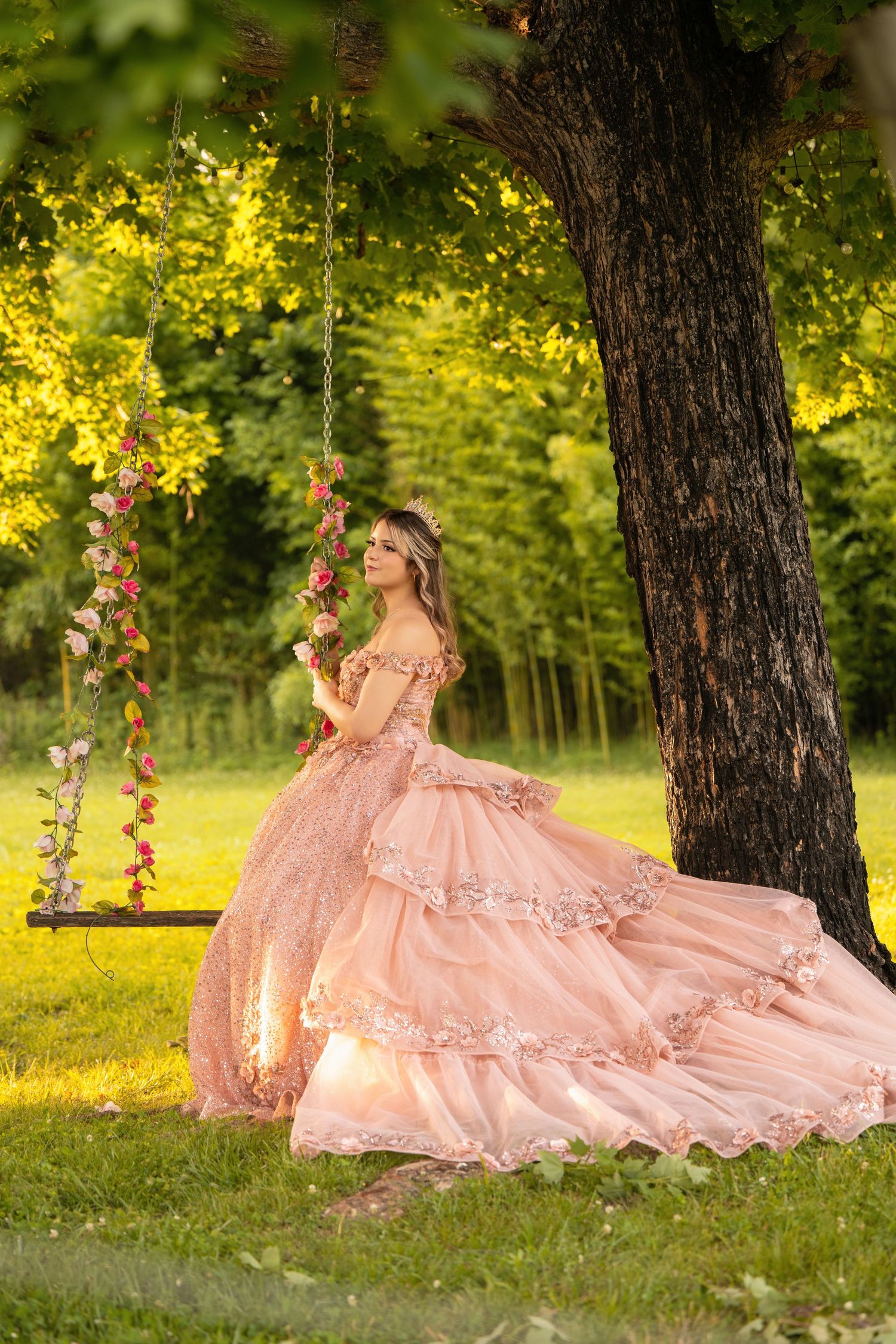 Young woman in elegant pink ball gown standing near a tree swing adorned with flowers in a lush garden.