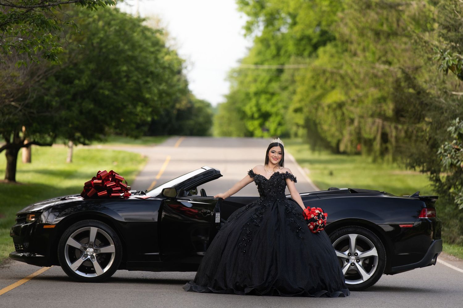 Young woman in black ball gown poses beside a black convertible sports car adorned with red bows on a tree-lined road.