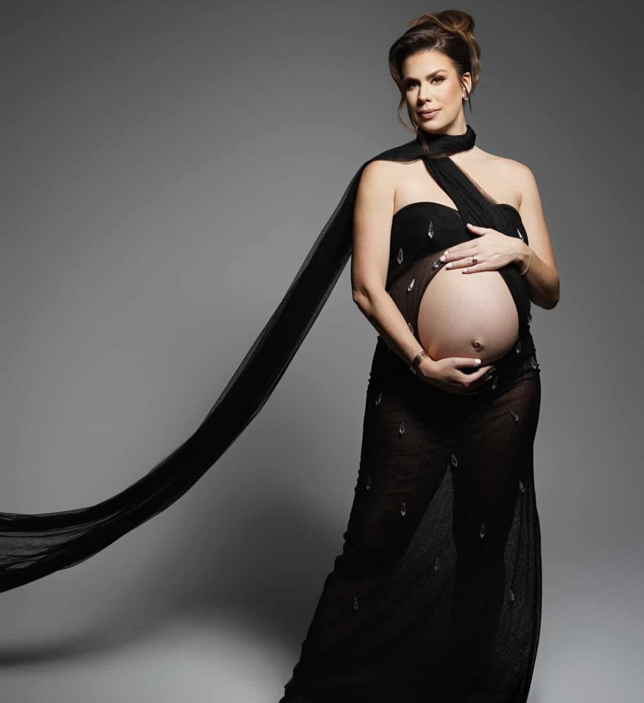 Pregnant woman in elegant black flowing maternity gown posing gracefully, showcasing baby bump in studio photo.