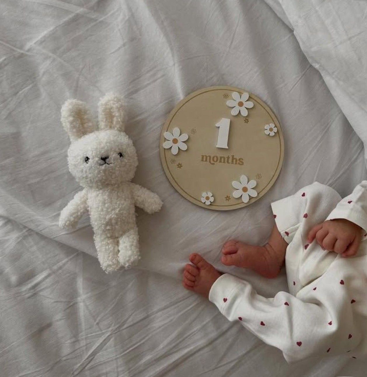 A 1-month-old baby sleeping on white bedding beside a plush bunny toy and a wooden milestone disc.