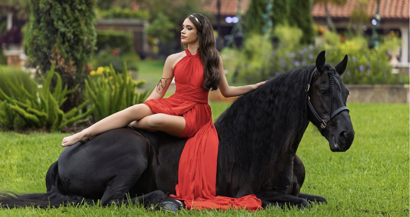 Woman in red dress posing elegantly with a black horse on green grass in a lush garden setting.