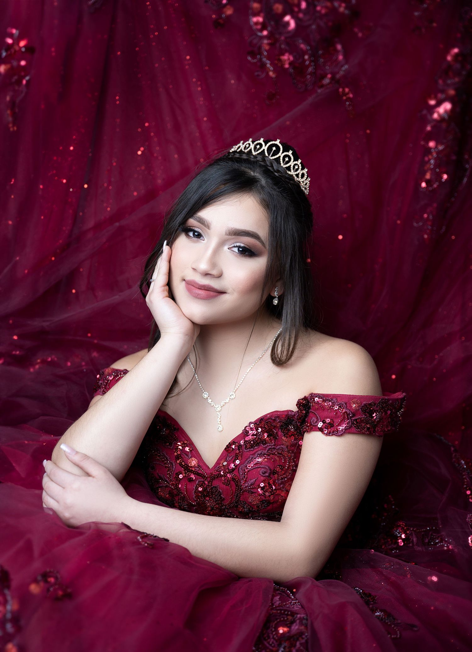 Elegant formal portrait in sparkling burgundy gown with delicate crown against red backdrop.