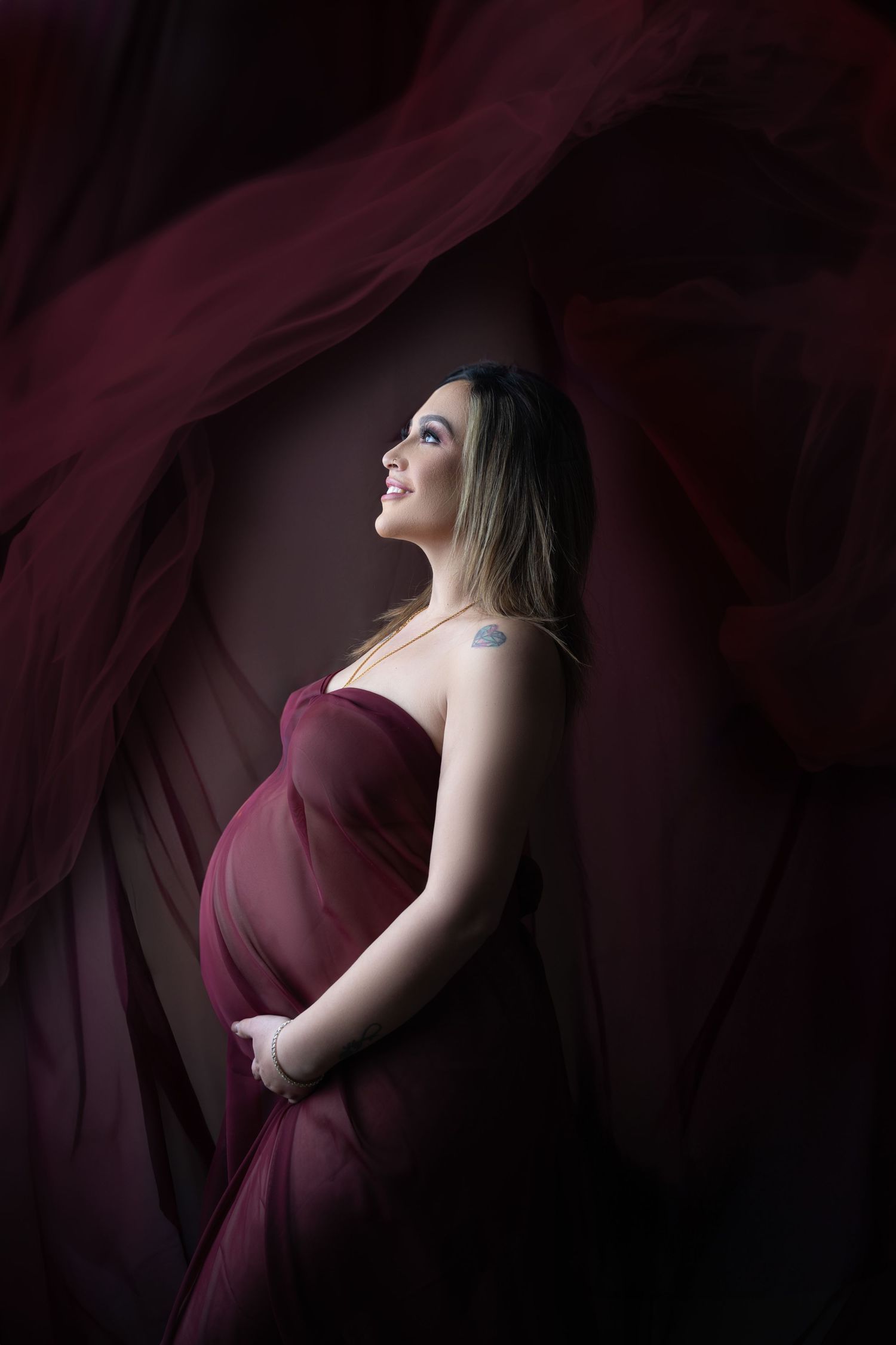 Dramatic maternity portrait with flowing burgundy fabric in dark setting.
