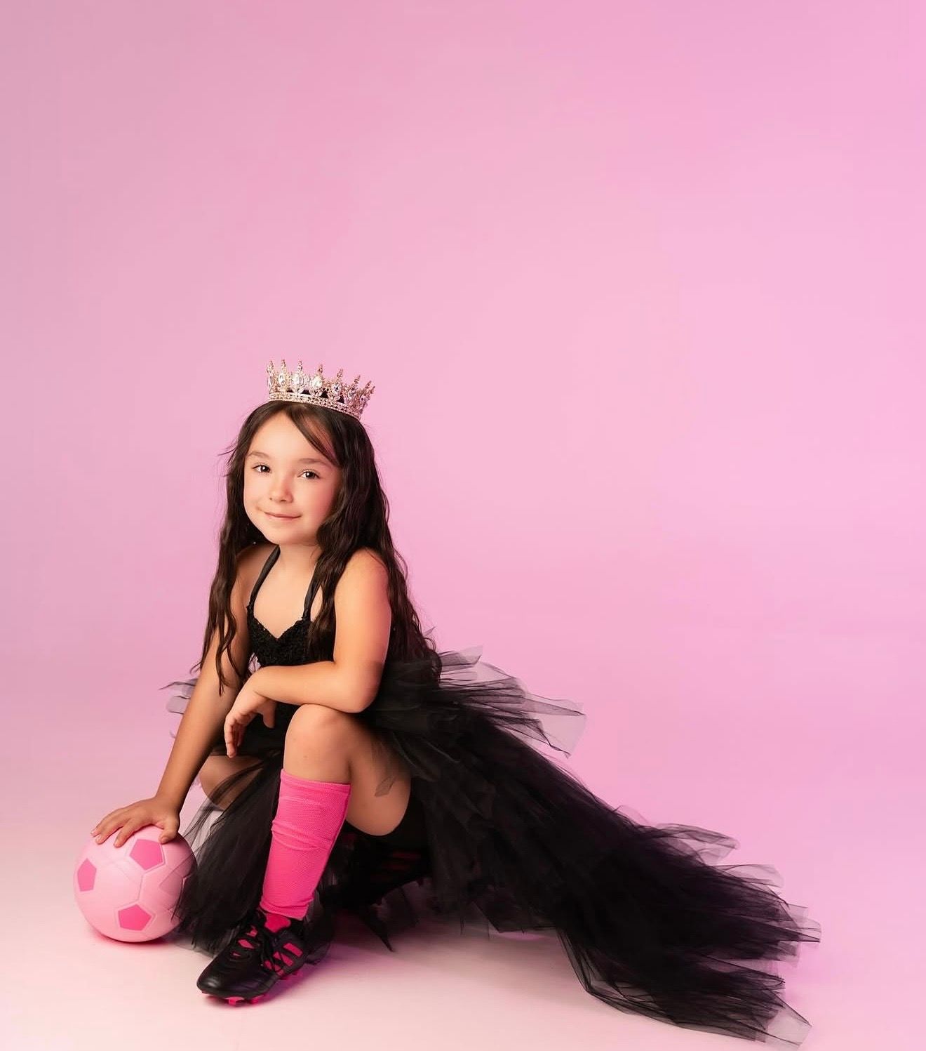Young girl in black tutu dress and crown sits on pink background holding a pink polka dot ball wearing pink knee socks.