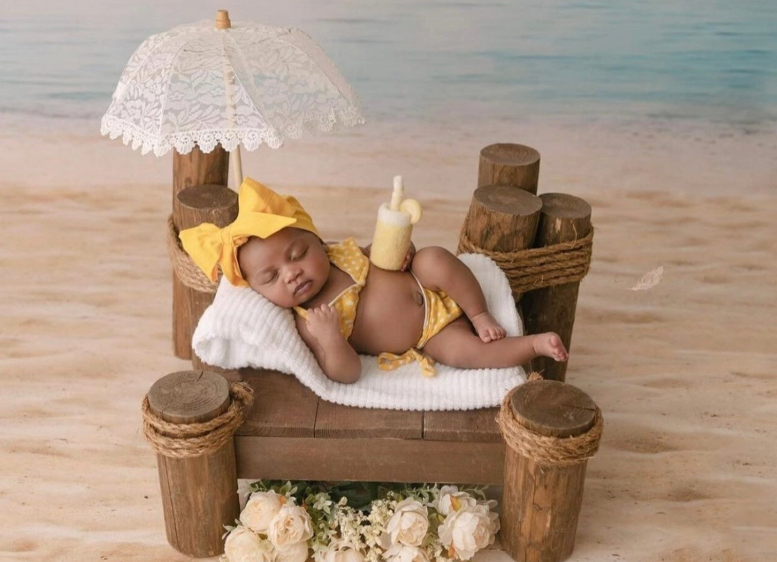 A baby in yellow sleeps on a wooden beach bed with a lace umbrella and milk bottle on a sandy backdrop.