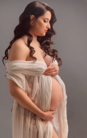 Elegant maternity portrait featuring a woman in flowing white fabric cradling her pregnant belly against a gray backdrop.