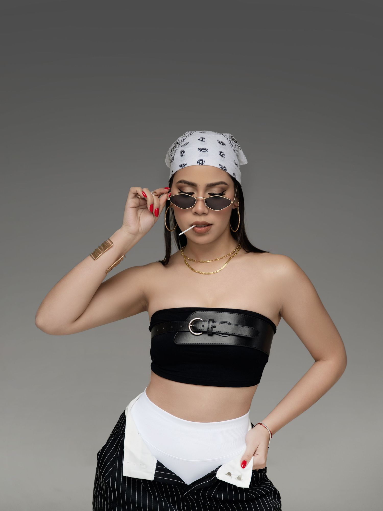 Woman in black tube top, white bandana, sunglasses, gold jewelry, and striped pants poses confidently.