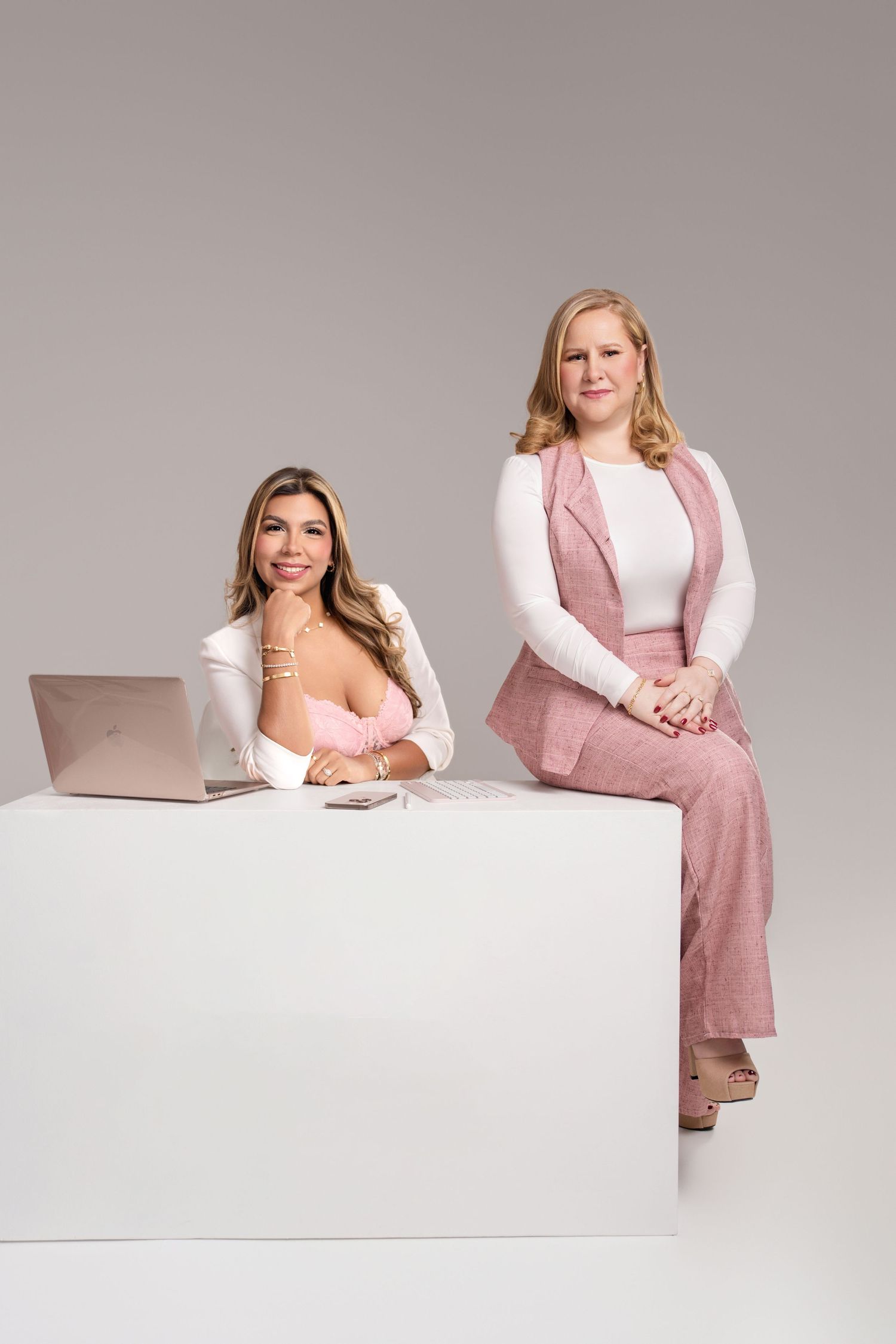 Two professional women in pink outfits pose confidently with a laptop at a white desk in a studio setting.