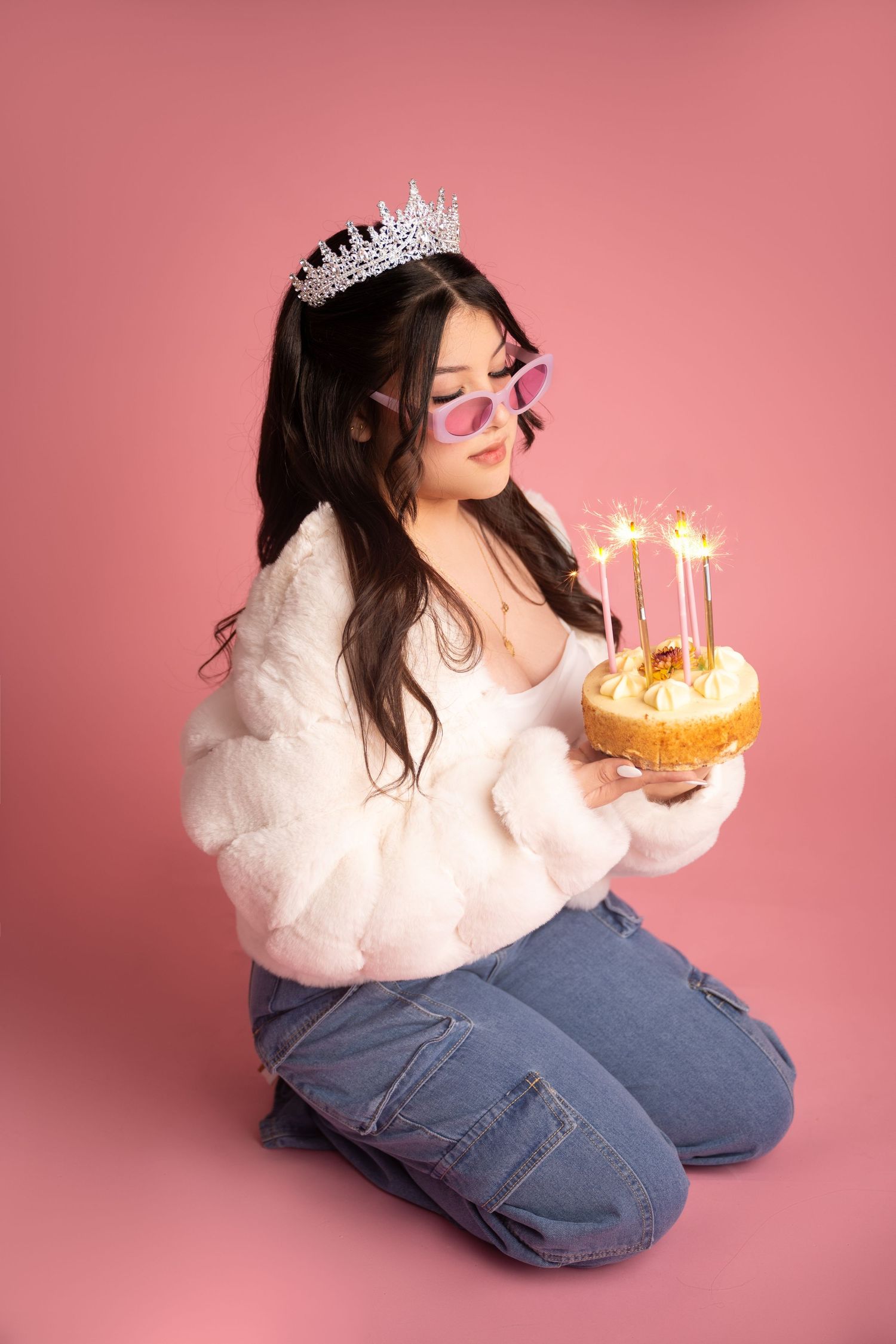 Young woman in fluffy white jacket, tiara, and pink sunglasses kneeling while holding a birthday cake with sparklers.