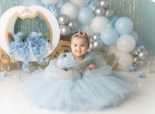 Baby girl in light blue tulle dress sits smiling surrounded by blue balloons and floral decorations for a princess-themed photo shoot.