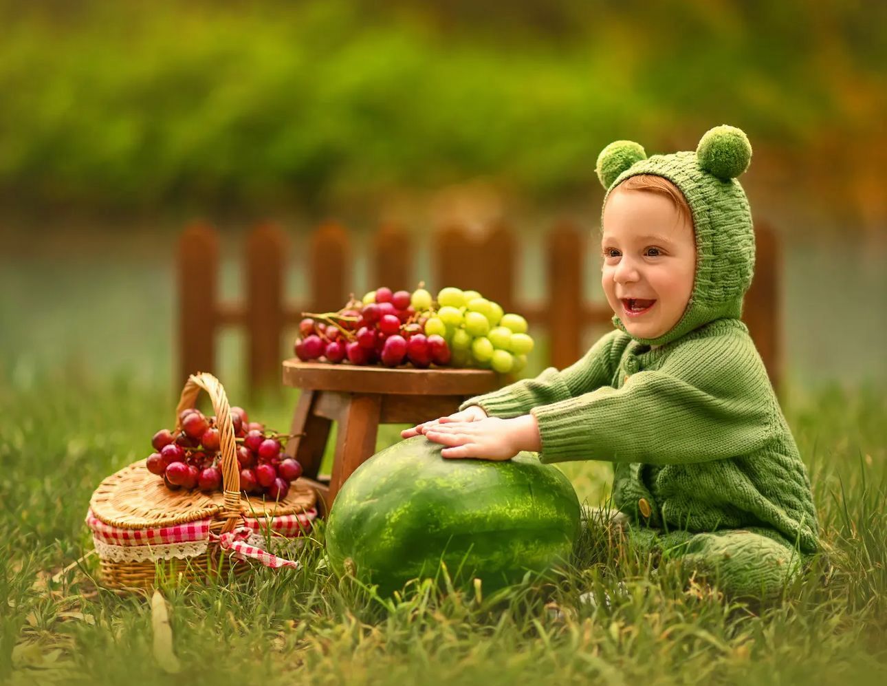 A cheerful baby in a green frog costume sits on grass beside a watermelon and basket of grapes at a tiny table.