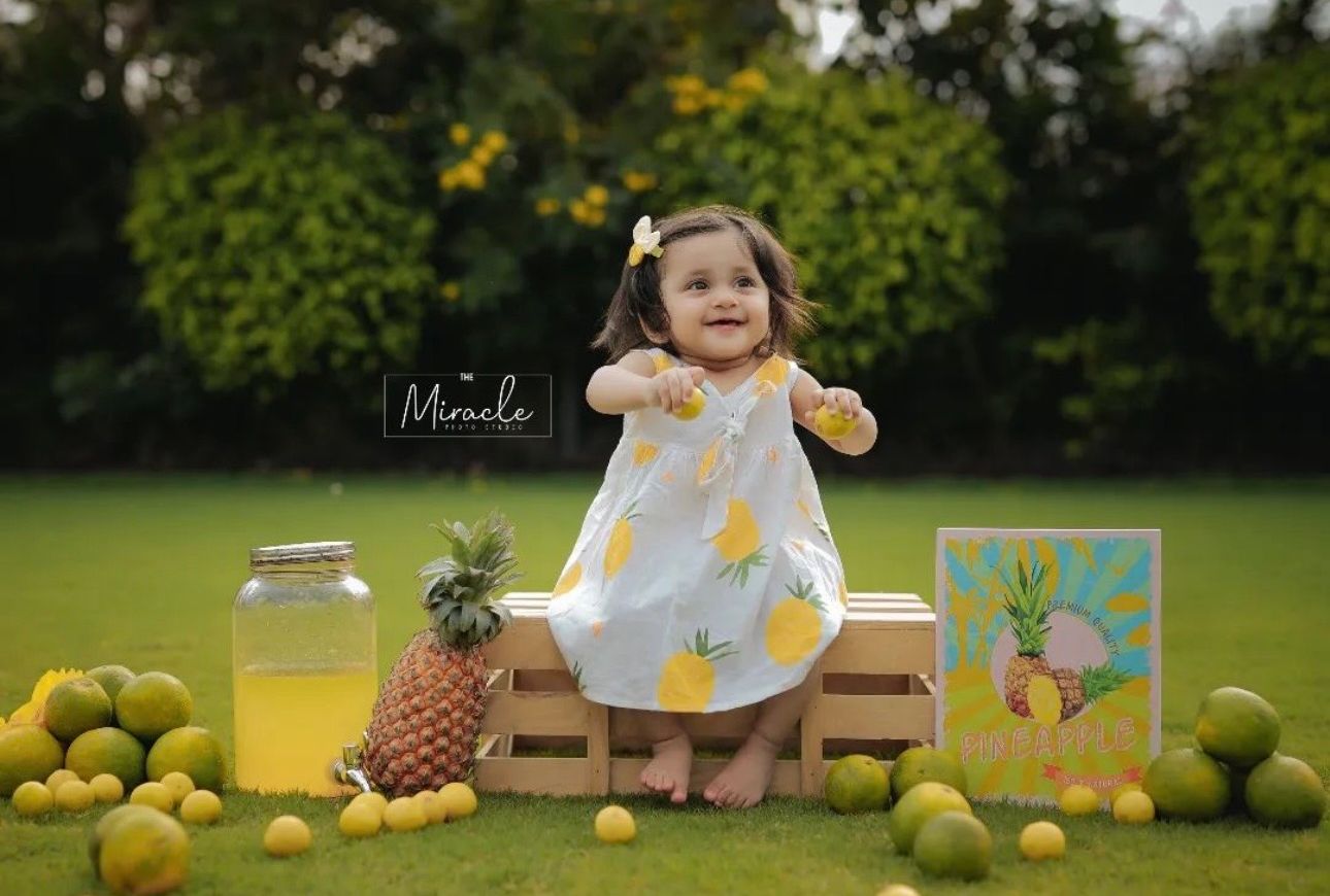 Adorable baby girl in pineapple dress sits on wooden crate surrounded by tropical fruits in lush green garden.