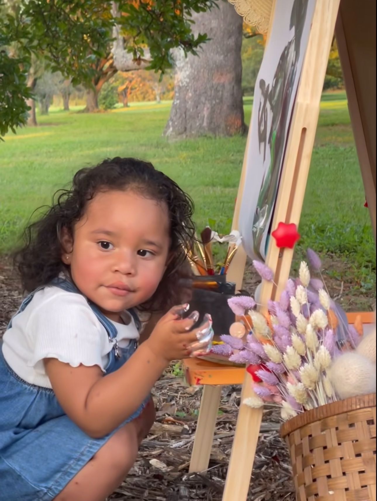 Toddler girl with curly hair in denim overalls painting at a wooden easel outdoors in a green park.