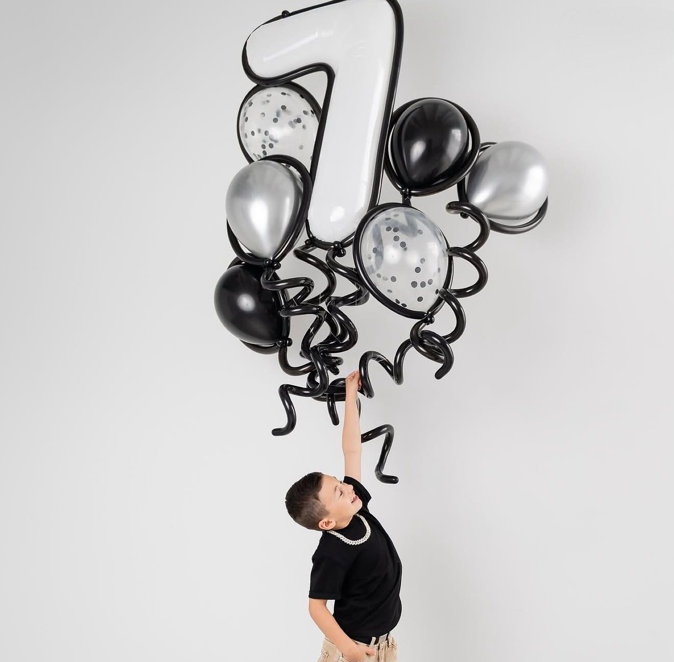 Boy reaching up to hold black, silver, and white number 7 balloon bouquet with curly ribbons for a 7th birthday.