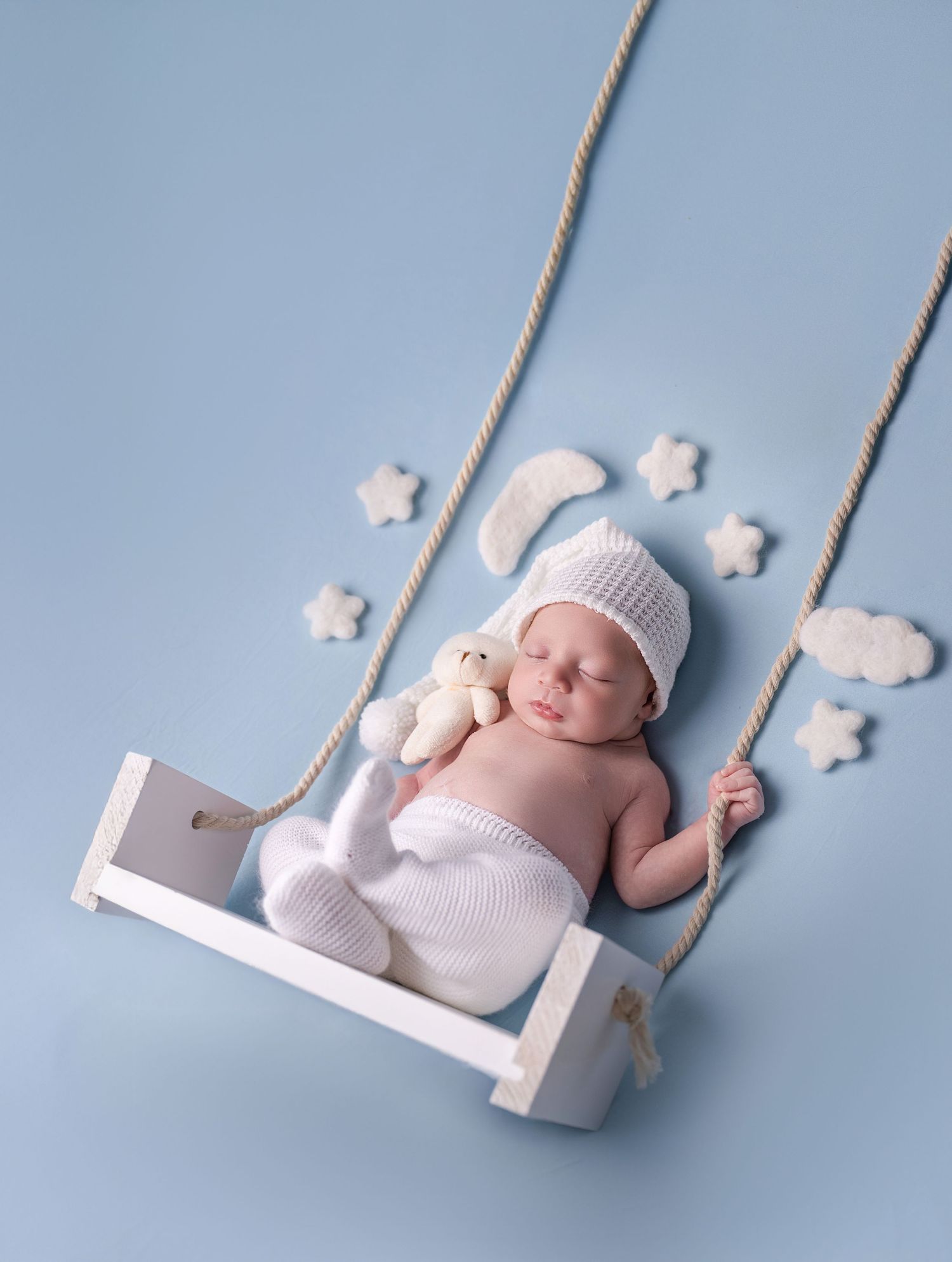 Sleeping newborn baby in white knit hat holds teddy bear on a small wooden swing with stars and clouds on blue background.