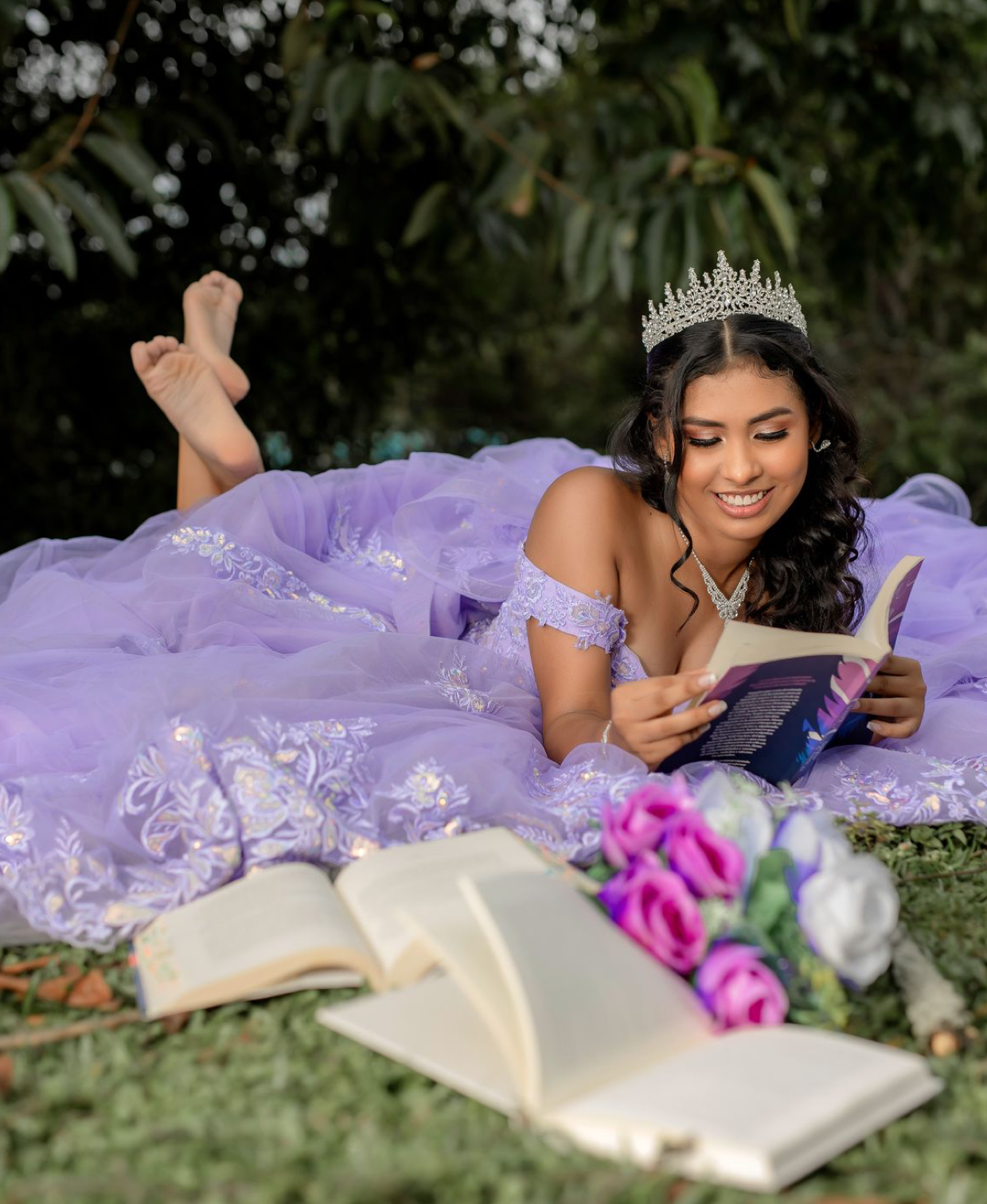 A young woman in a lavender quinceañera gown and tiara reads a book outdoors surrounded by flowers.