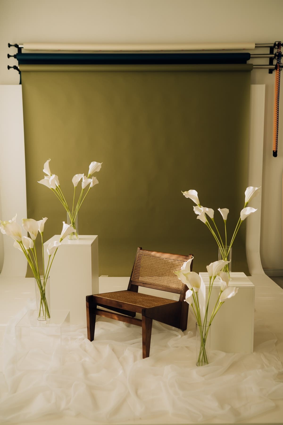 Minimalist studio setup with white calla lilies in vases, a wooden chair, and an olive green backdrop.