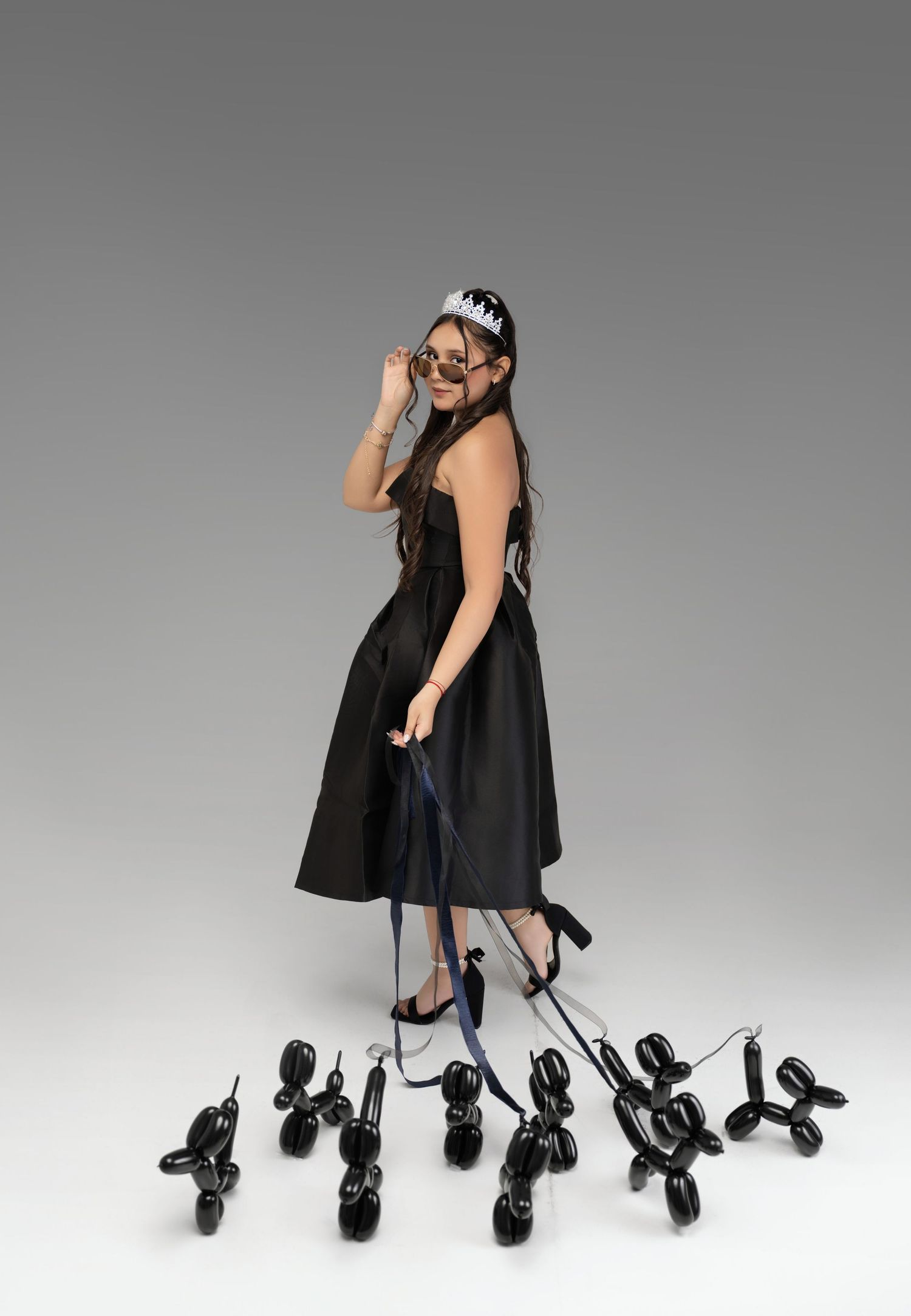 Woman in black dress and crown holds leashes of black balloon dogs while adjusting sunglasses.